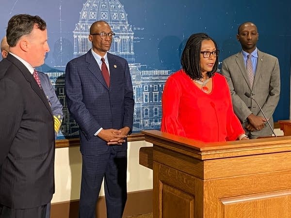 A woman stands behind a podium, four men stand around her listening.