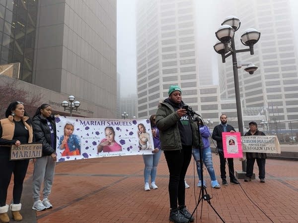 A person speaks at a mic while others hold a banner for Mariah Samuels in the background.