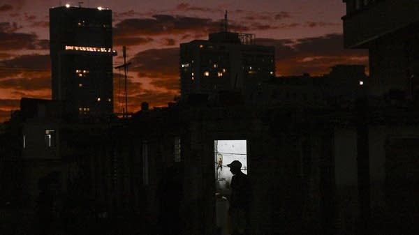 The silhouette of a man is seen at his home during a blackout in Havana in front of a sunset and tall, darkened buildings.