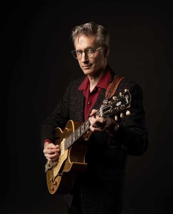A man in a suit poses for a photo while playing guitar