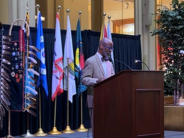 A man speaks at a podium in front of various flags.