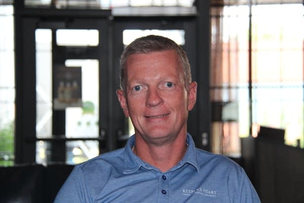 A man wearing a blue polo shirt looks directly into the camera. He sits in a restaurant.
