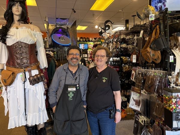 A man and woman pose in their costume shop next to a mannequin in renaissance garb.