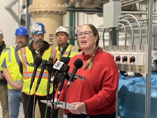 A woman stands in front of a stand of microphones. Large water pipes are behind her.
