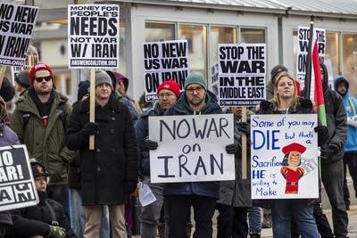 A crowd stands outside holding signs to protest military action in Iran.