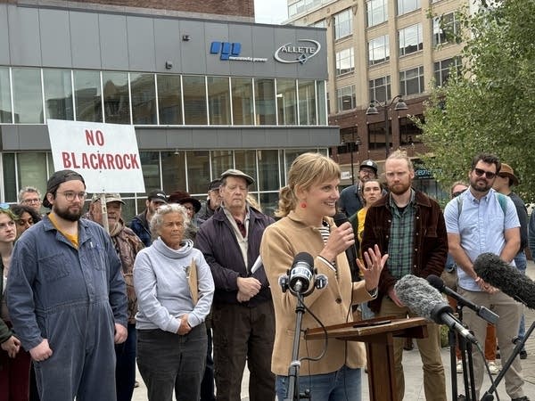 A woman speaks at a podium. Behind her is a crowd of protesters standing in front of the Minnesota Power headquarters.