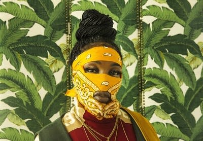 A woman with a bandana face covering in front of fern-patterned wallpaper