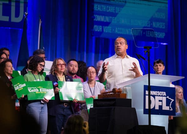 Man in white shirt standing at podium with people holding signs behind him