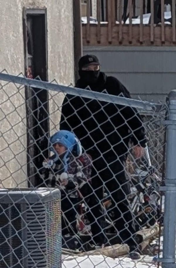 A masked officer stands behind a young boy behind a chain link fence.