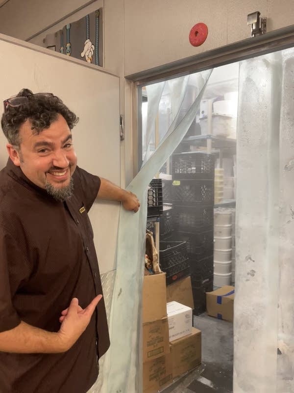A man in a dark shirt and curly hair holds open the curtain to a walk-in freezer. 