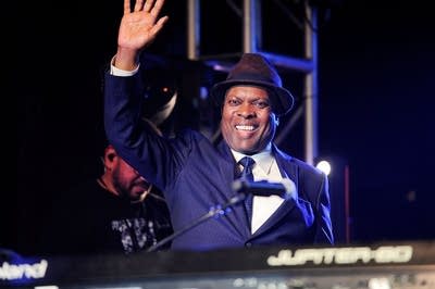 A man seated at a piano onstage smiles and waves
