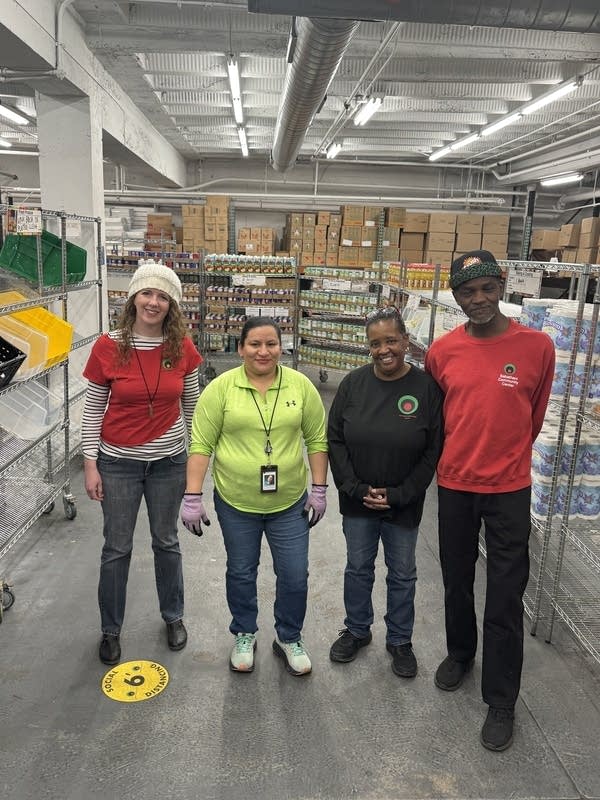 Four people stand in a warehouse with food on shelves.