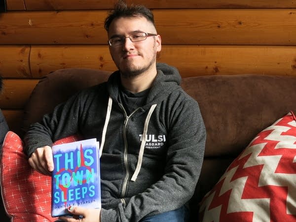 A man sitting in a rustic setting holds a novel to display the cover.