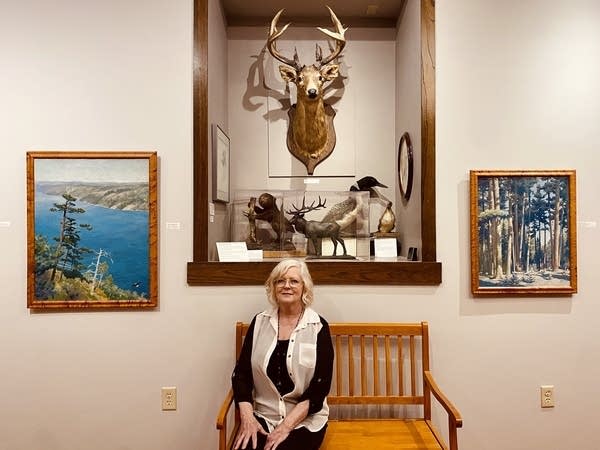 A woman sits on a bench with taxidermy behind her and paintings hung on either side.