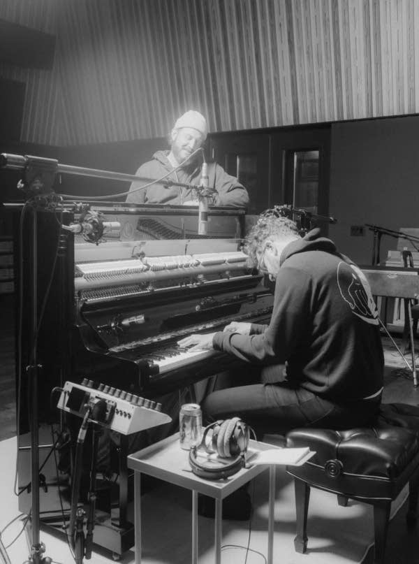 In a recording studio, one man plays piano while another stands against it, listening