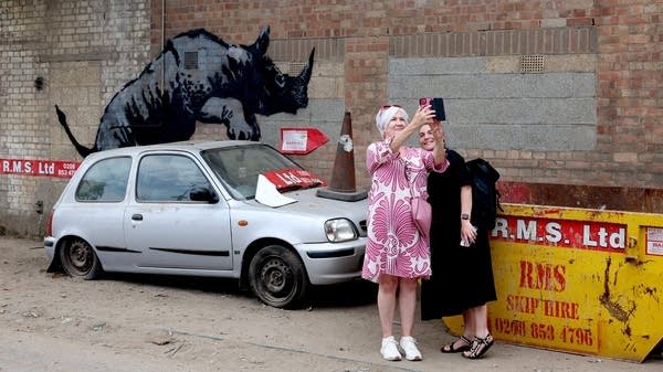 Two people take a selfie in front of a Bansky piece of a rhino mounting a car with a flat tire.