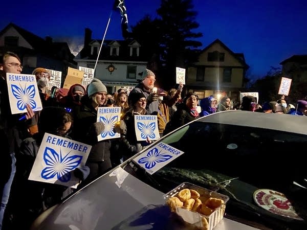 A group gathers around a car, holding signs that read "Remember" with a blue butterfly.