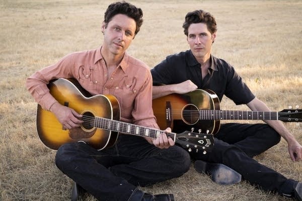 Two men sit in field holding acoustic guitars and looking at camera.