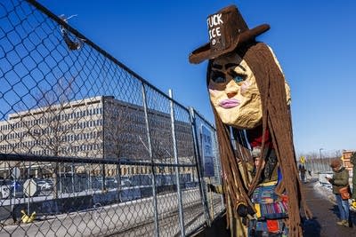 A protester holds an oversized puppet head featuring long brown hair, large eyelashes and a cowboy hat that reads "F--- ICE."