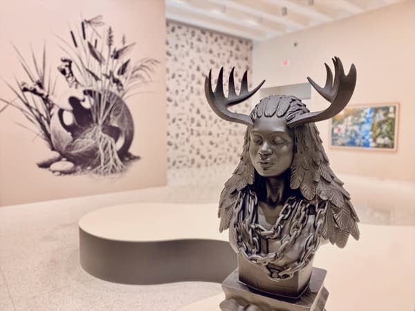 A gray, 3D-printed sculpture of a woman with antlers is displayed in a museum.