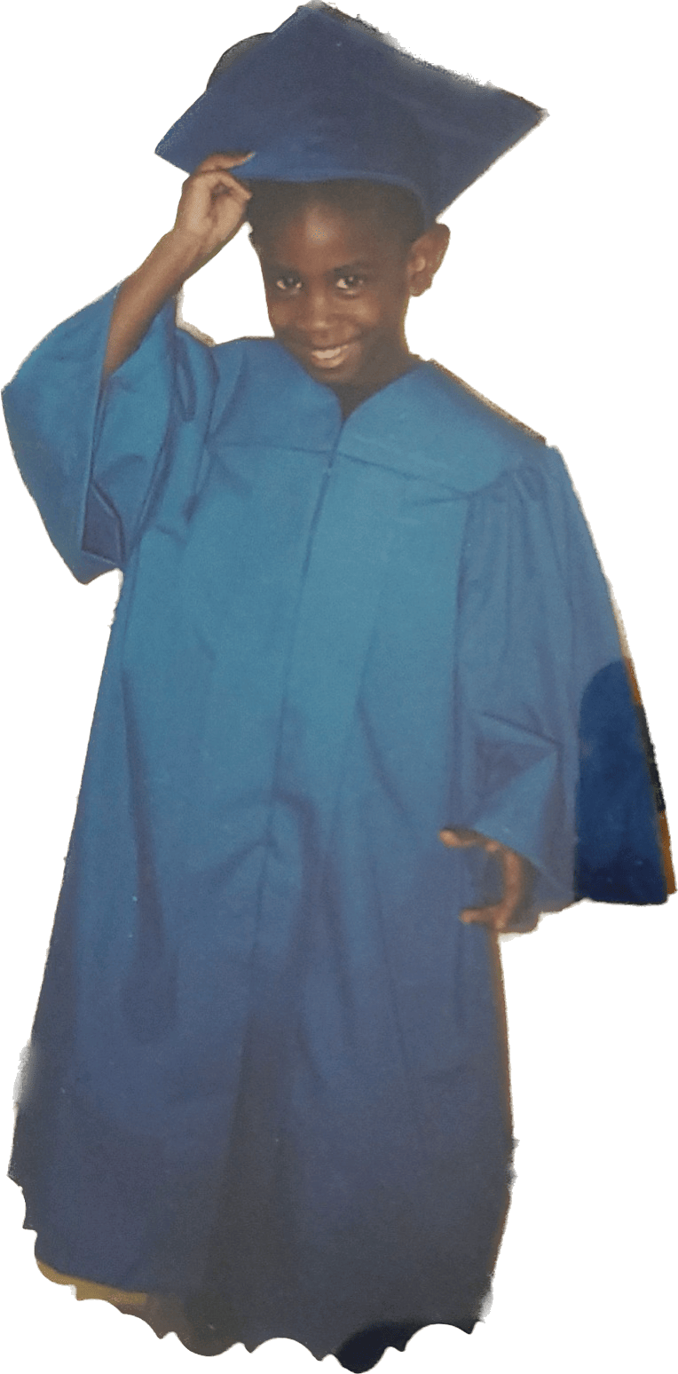 A young Black boy stands wearing a blue graduation cap and gown. He is smiling at the camera and holding his cap.