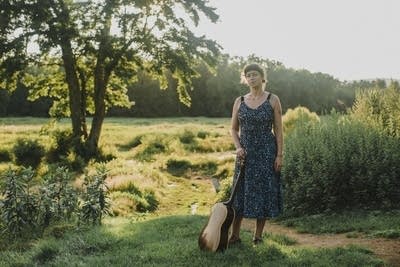A woman with a guitar stands in a sunny meadow