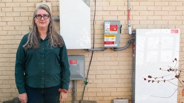 A woman in a green shirt stands next to a brick wall with a solar and battery power system.