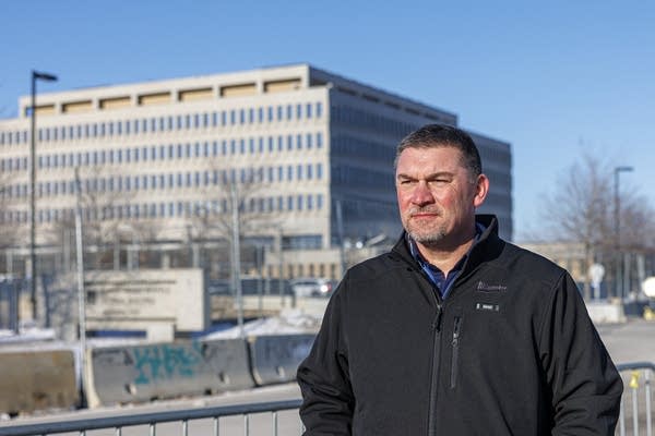 A man in a black jacket stands with a large gray concrete building behind him.