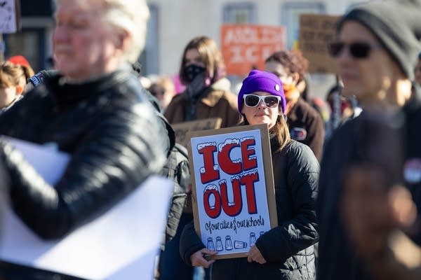 A woman in a crowd holds an "ICE Out" protest sign.