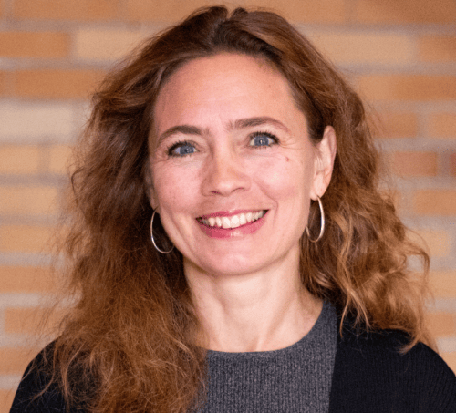 Kristin Persson poses for her headshot in front of a red brick background wearing a grey and black top and smiling.