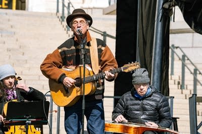 Three musicians perform together on an outdoor stage at a large rally
