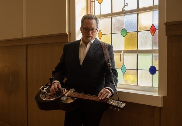 A man plays dobro while standing next to a leaded and stained glass window