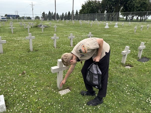 A boy scout leans down to clear off a grave in a cemetery.
