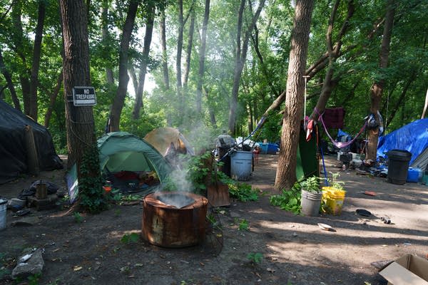 Wide shot of an encampment site in a clearing in the woods. Smoke rises from a fire pit in the center.