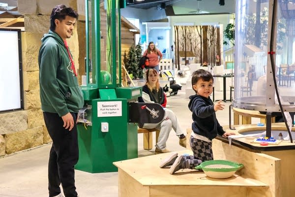 A man stands and watches as a child plays with an exhibit at the museum.