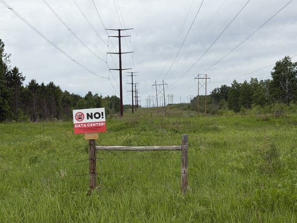 A red and white sign reading "NO! Data center" sits on a wooden fence post in a clearing under a power line.