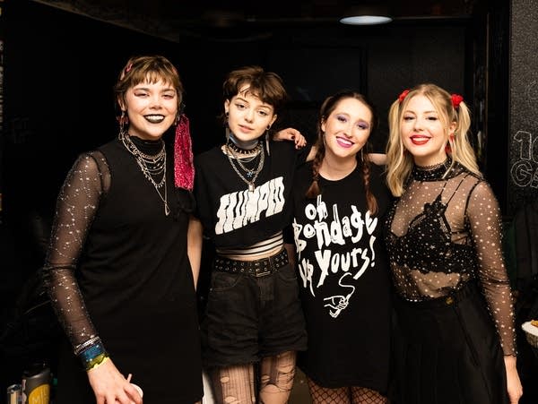 Four band members stand with their arms around each other's shoulders