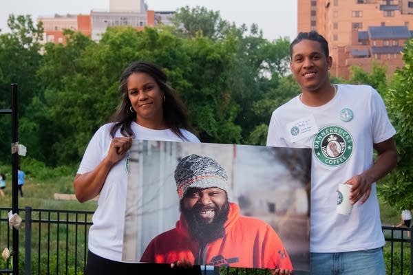 Two people stand holding a large photo of a man. 