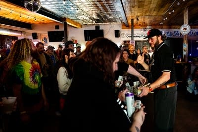 Justin Vernon wears a black shirt and cap at a bar and hands out pull tabs.