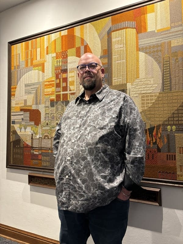 A man in a gray shirt poses in front of a large painting.