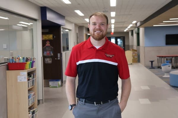 A man in a red and black polo smiles for a photo in an empty school hallway.