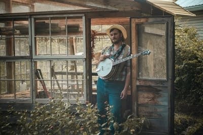 A man stands outside a potting shed, holding a banjo 