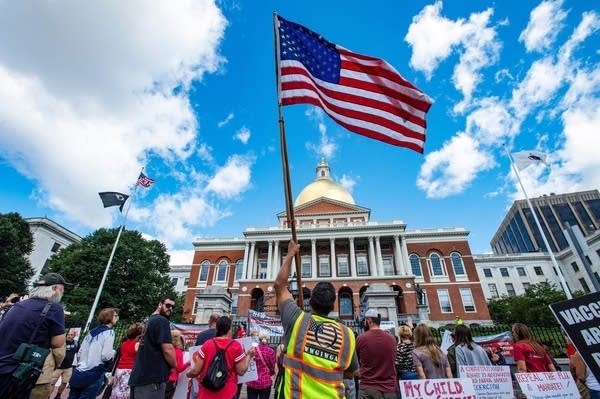 A man with a QAnon vest and U.S. Flag joins hundreds of people protesting a mandate from the Massachusetts governor requiring all children to receive a flu vaccine in order to attend school.