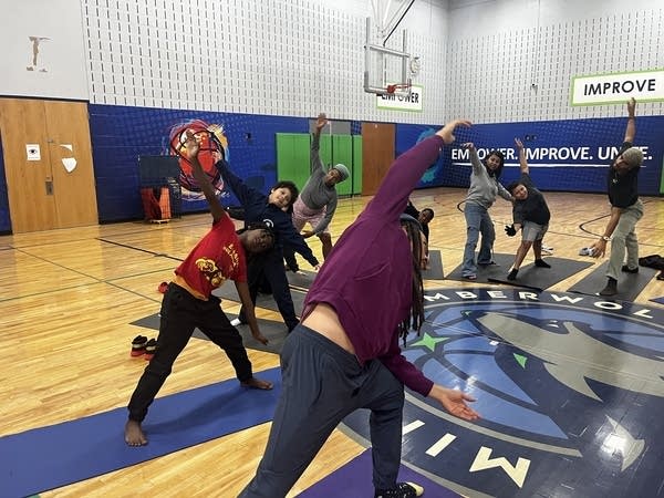 Kids stretch their arms over their heads while on yoga mats in a gymnasium. 