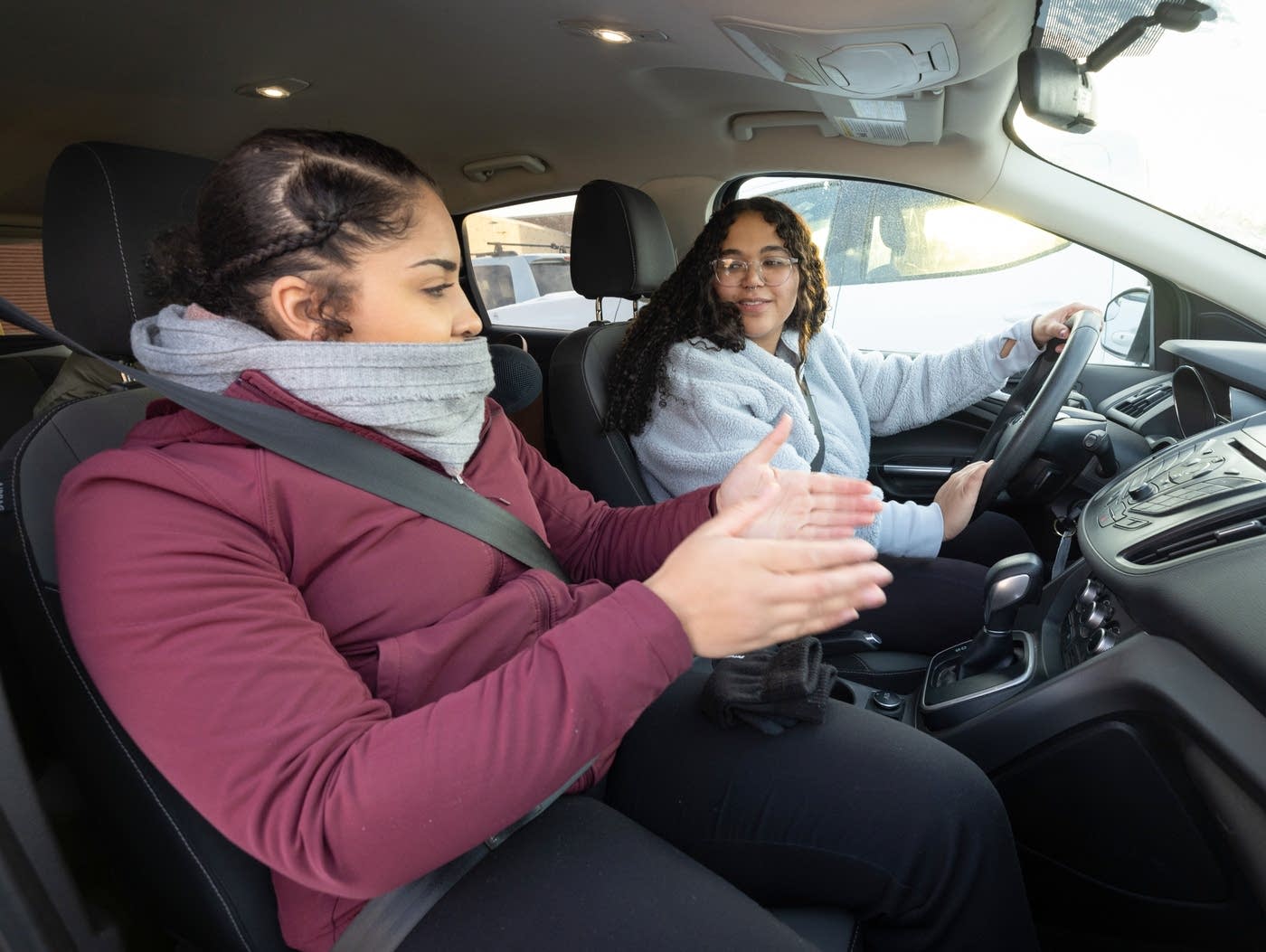 Two people sit in a car, one holds out two hands as she talks to the other behind the wheel.