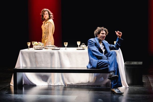 A woman stands behind a table while a man sits in front, on a theatrical stage.