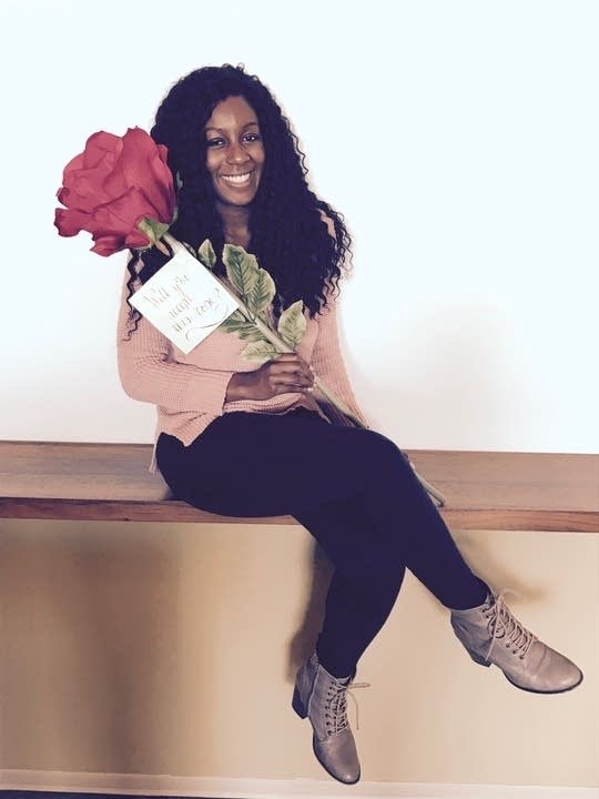Jazzy Collins, a Black woman with long hair wearing a pink cardigan, black pants and a blue sits on a wooden ledge. Her legs are crossed and she is holding a massive rose.