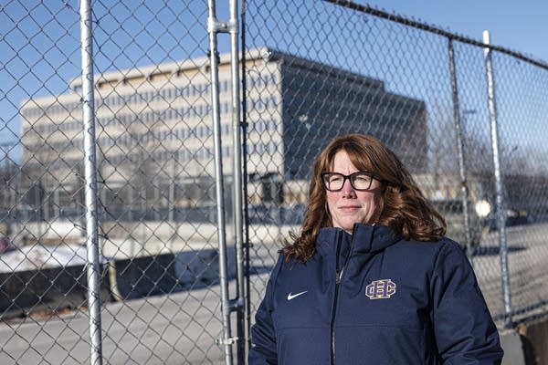 A woman in a navy coat stands in front of a fence enclosing a large, gray concrete building.