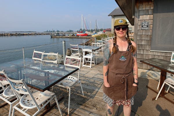 A woman in a brown apron poses for a photo outside a cafe overlooking a lake.
