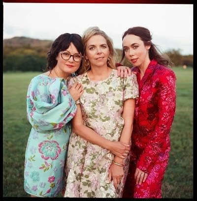 Three women in summer outfits stand outdoors on a grassy field